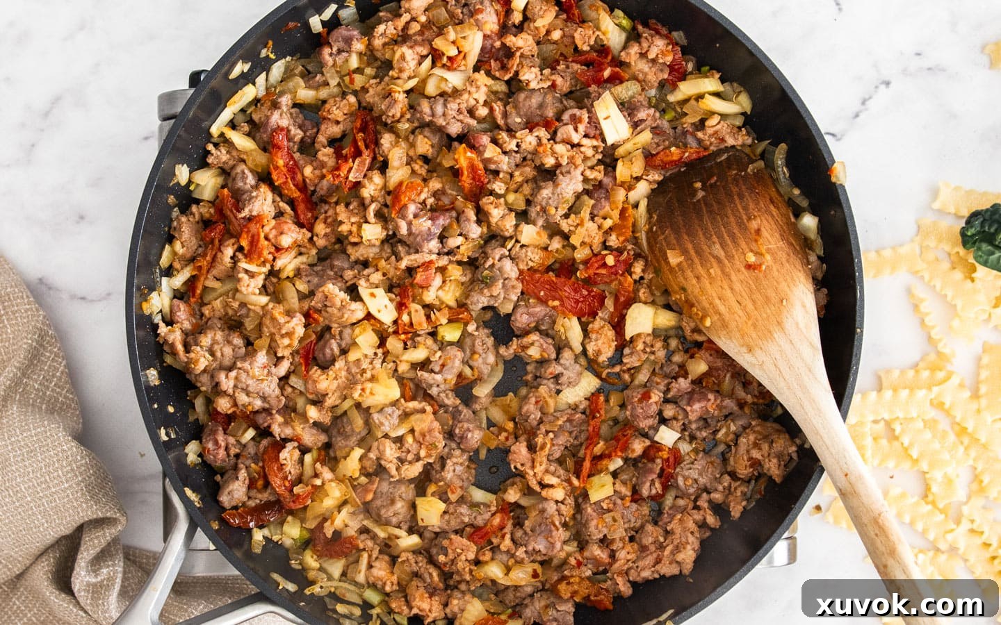 Crumbled Italian sausage cooking and browning in a pan with the sautéed vegetables.