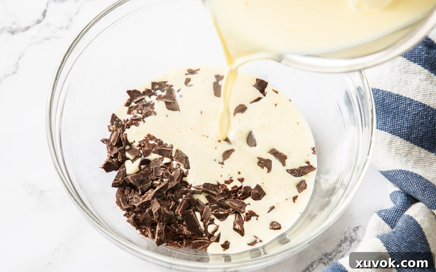 Hot cream being poured over finely chopped dark chocolate in a bowl to create the chocolate cream base.