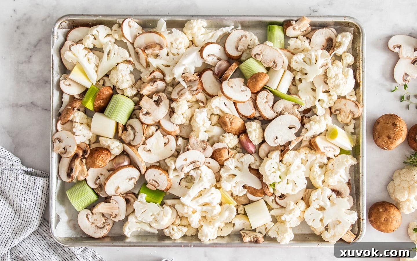 Cauliflower, mushrooms, leeks, and unpeeled garlic cloves on a baking sheet, drizzled with olive oil and seasoned, ready for roasting.