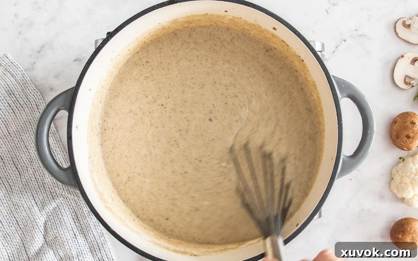 A whisk stirring creme fraiche into the blended mushroom cauliflower soup in a Dutch oven, making it extra creamy.