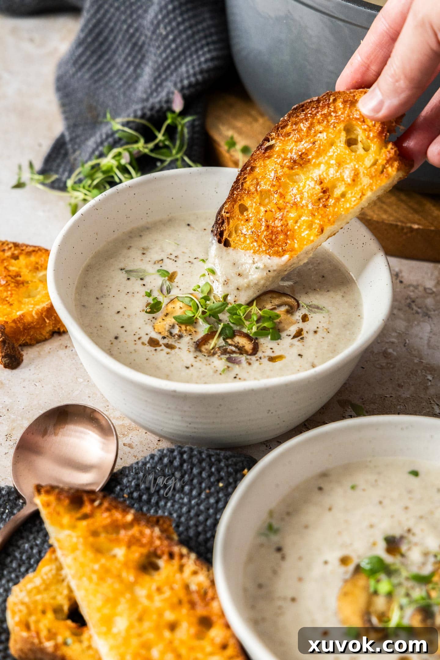 A golden, cheesy slice of bread being dipped into a steaming bowl of creamy mushroom cauliflower soup.