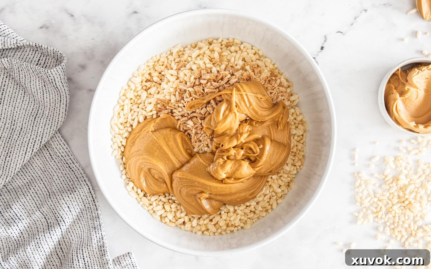 The smooth peanut butter mixture being poured into a large mixing bowl containing puffed rice cereal and chopped almonds.