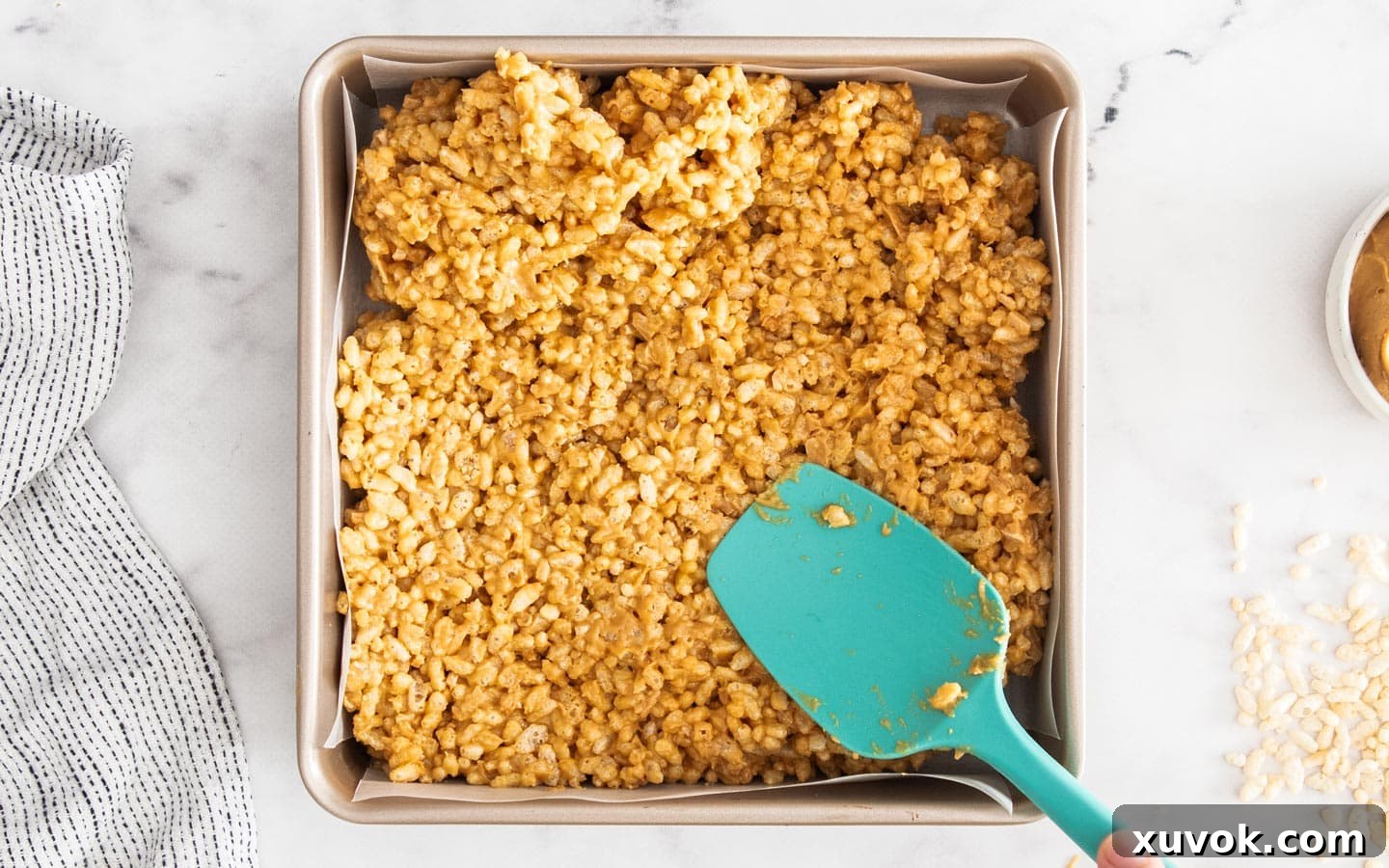 A spatula pressing the peanut butter puffed rice mixture firmly into a lined baking pan to create an even, compact base.