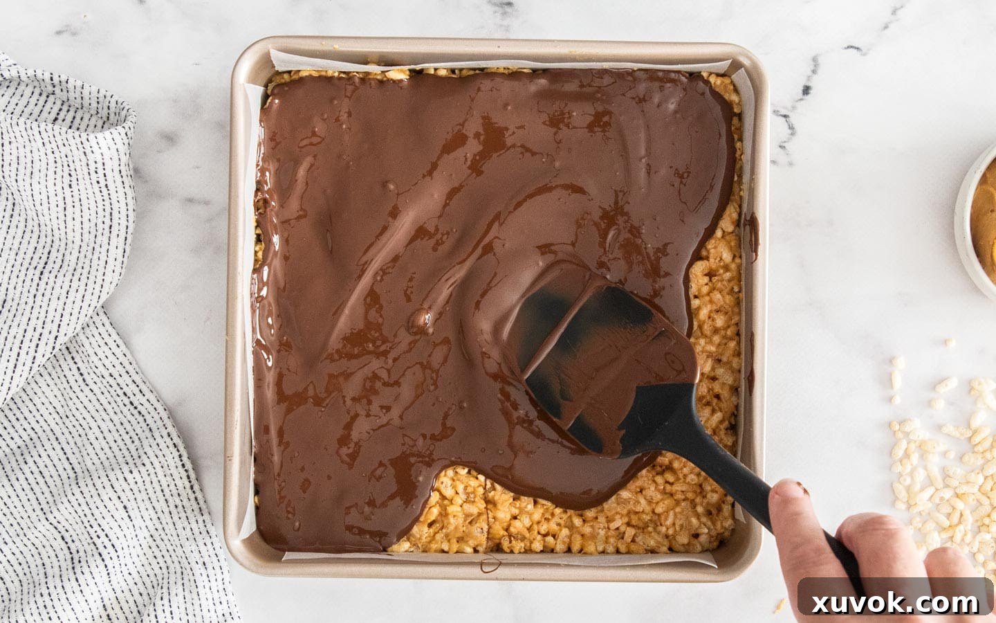 Melted chocolate being smoothly spread over the pressed peanut butter rice crispy base with a spatula.