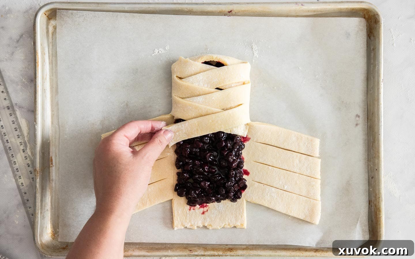 A hand gently pulling the sixth strip across the blueberry Danish braid, illustrating the alternating fold pattern.