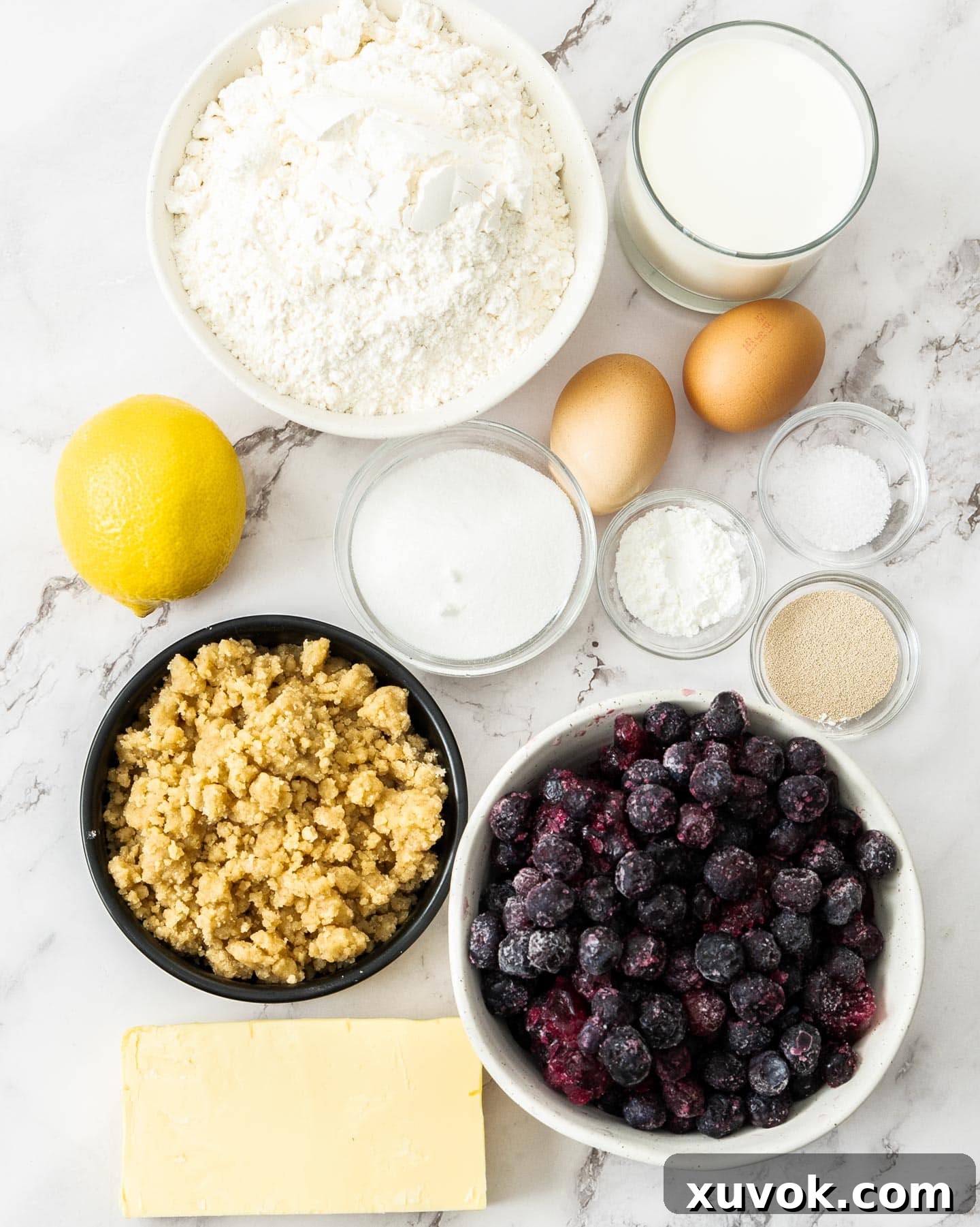A collection of fresh ingredients laid out for making blueberry Danish braid, including blueberries, butter, flour, eggs, and lemon.