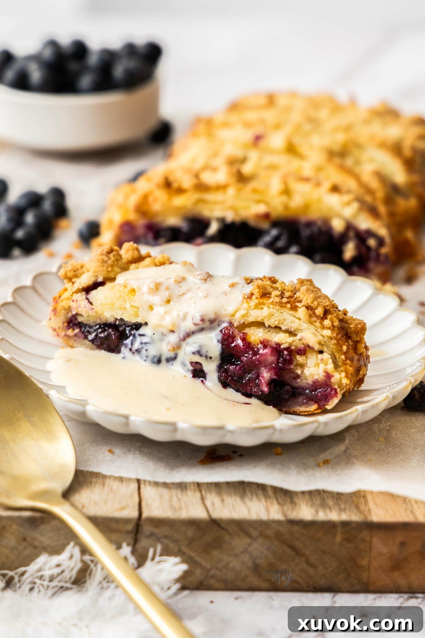 A perfectly sliced piece of blueberry Danish braid elegantly presented on a dessert plate, generously drizzled with rich crème anglaise.