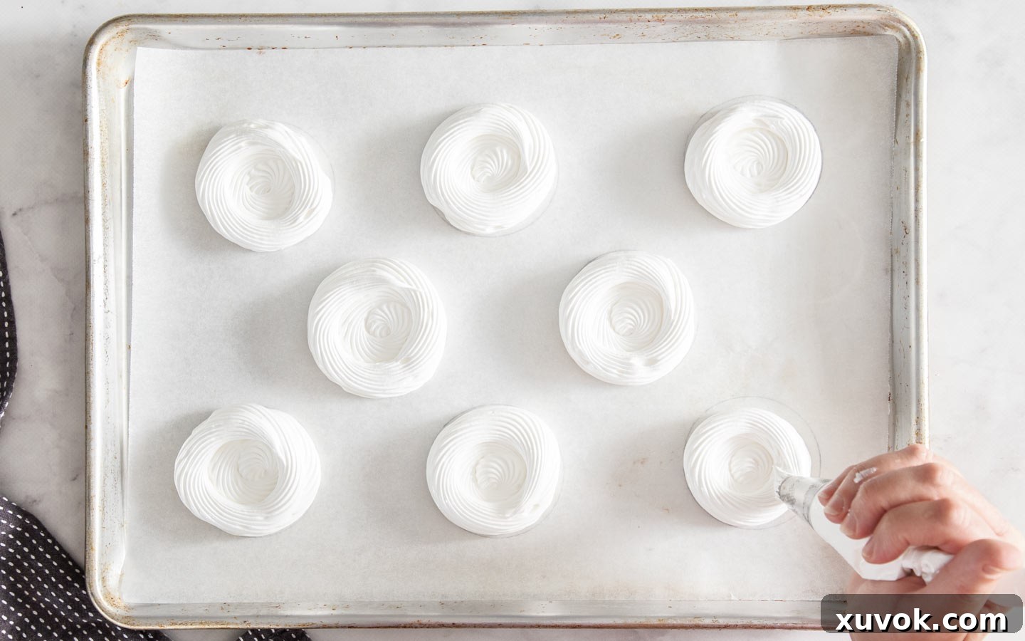 Hands piping spirals of meringue mixture onto baking paper, following circular guides, forming the base of the nests.
