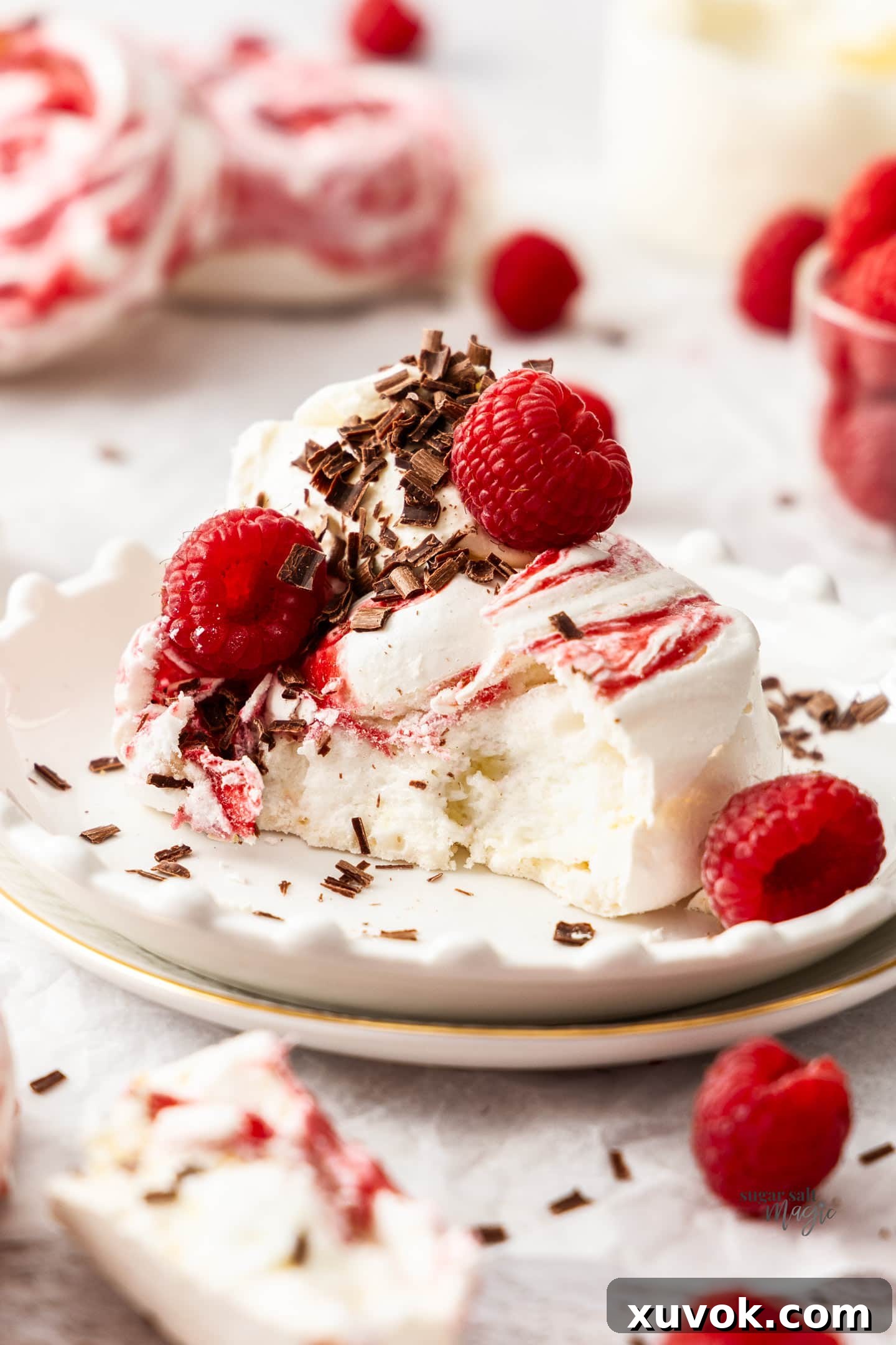 A beautifully cut raspberry meringue on a plate, topped with cream, fresh raspberries, and chocolate shavings.