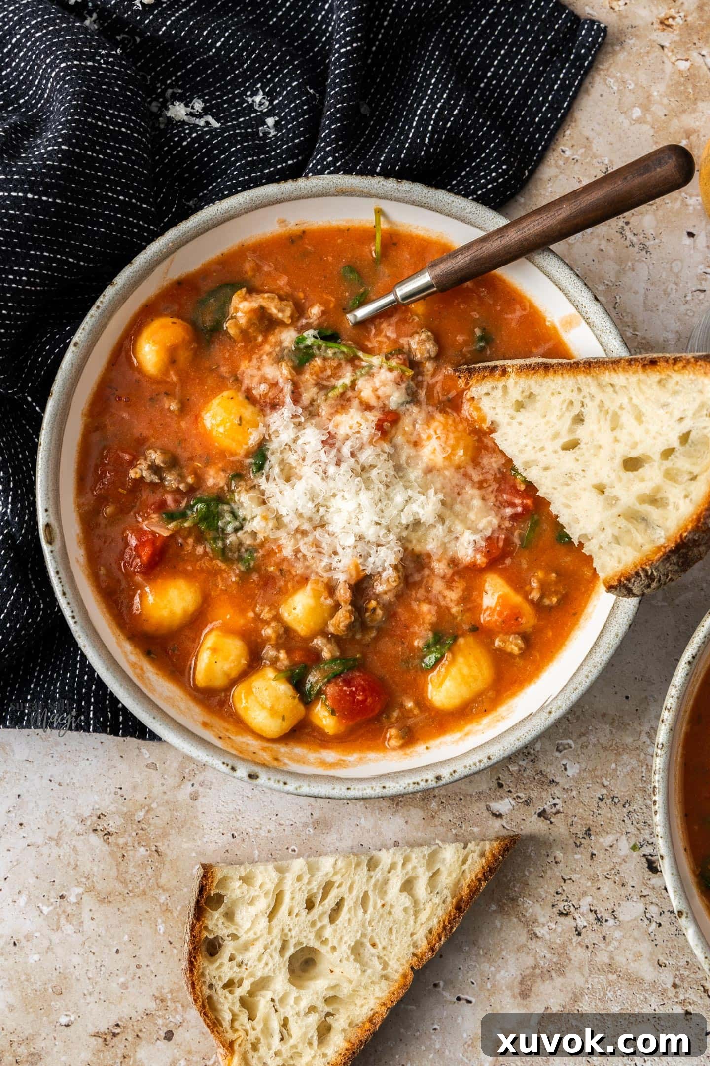 A top-down view of a steaming bowl of homemade sausage gnocchi soup, garnished with Parmesan, with crusty bread chunks on the side.