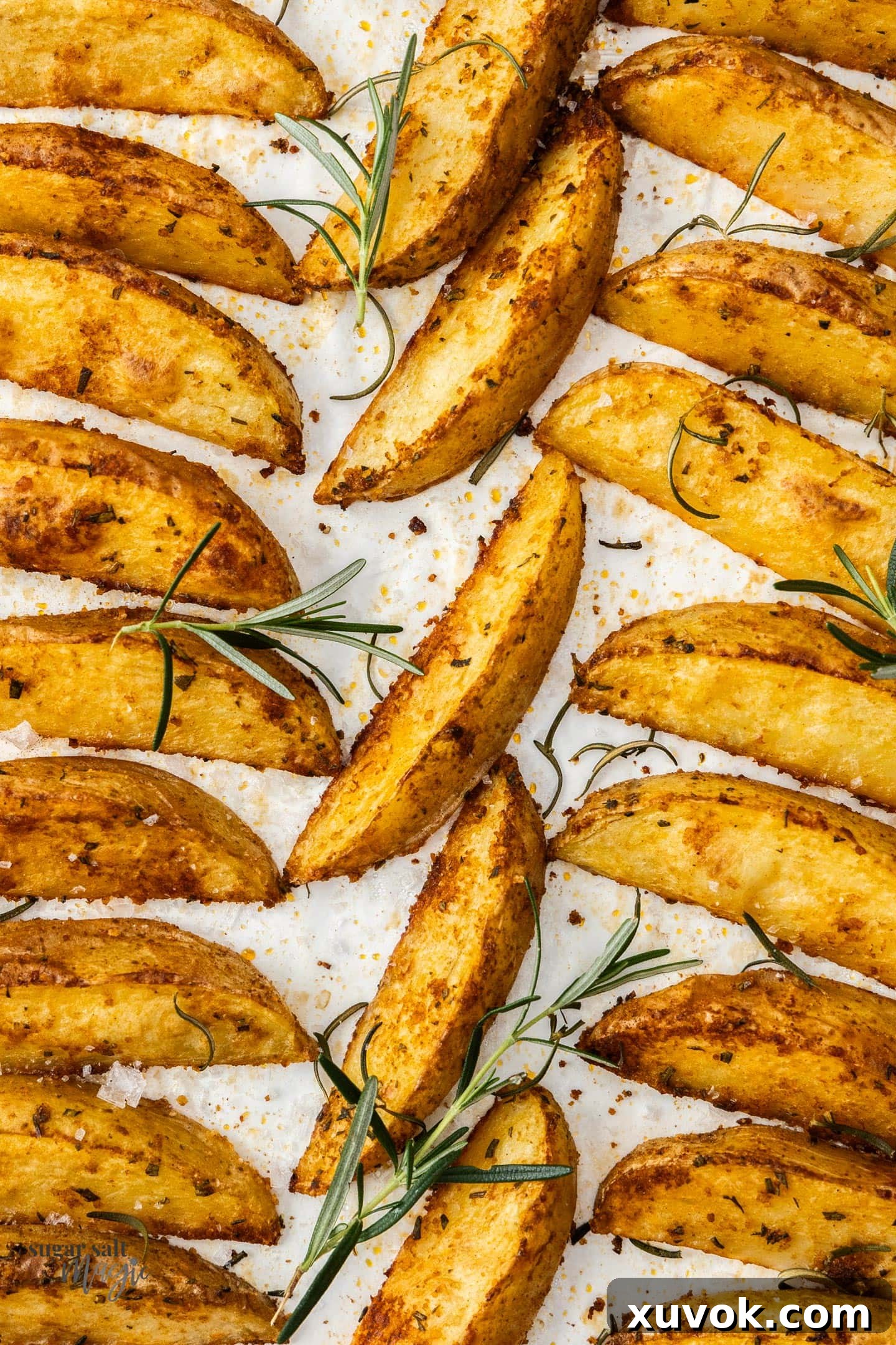 Top down view of freshly baked potato wedges on a baking tray, garnished with scattered fresh rosemary.