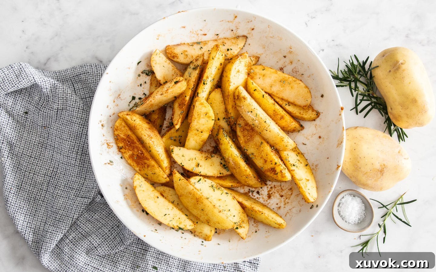 Unbaked potato wedges coated in seasoning and fresh rosemary in a large mixing bowl.