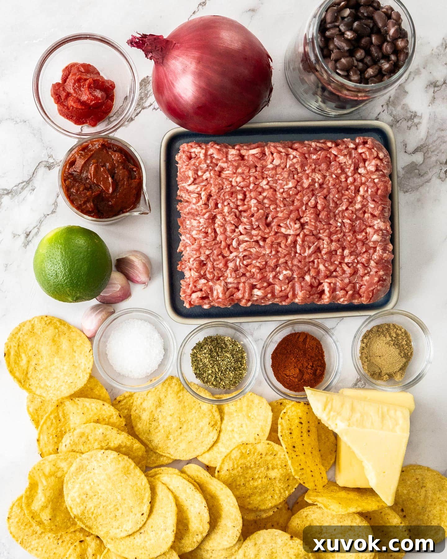 A selection of fresh ingredients laid out on a marble surface, including ground beef, corn chips, onions, garlic, spices, and a can of chipotle in adobo.