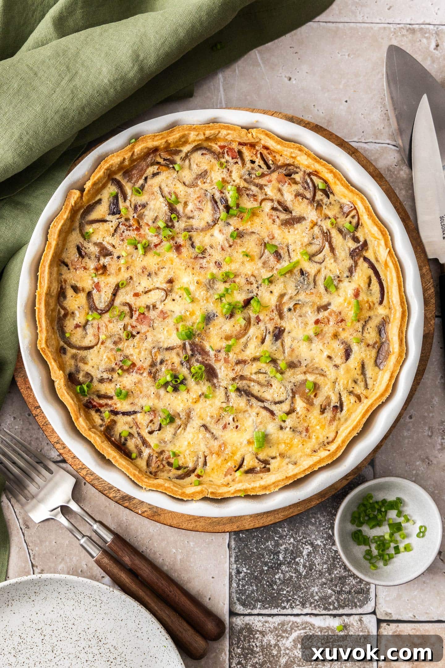 Top-down view of a golden, freshly baked Bacon and Caramelised Onion Quiche in a ceramic quiche dish, ready to be served. Forks and plates are neatly arranged to the side, hinting at a delightful meal.