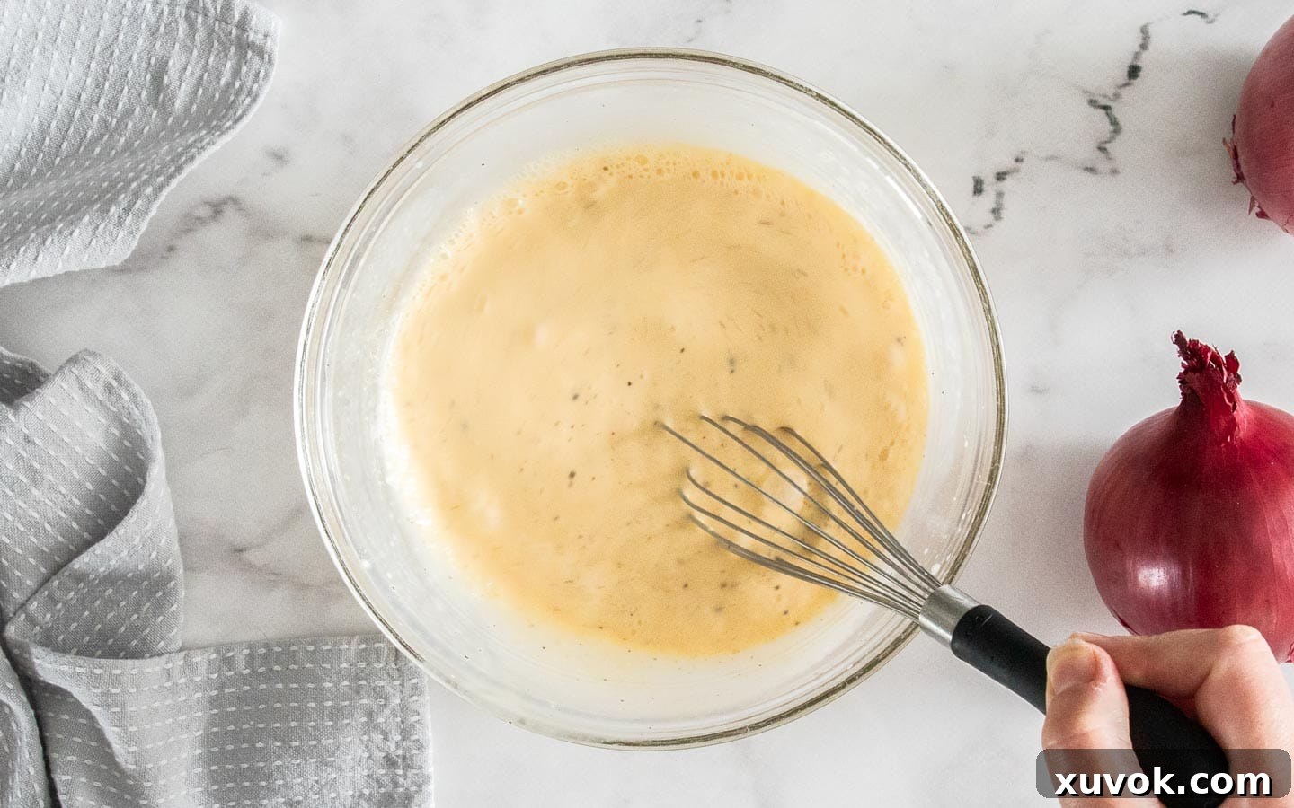 A hand whisking eggs, cream, salt, and pepper in a mixing bowl to create the smooth, creamy filling for the quiche.