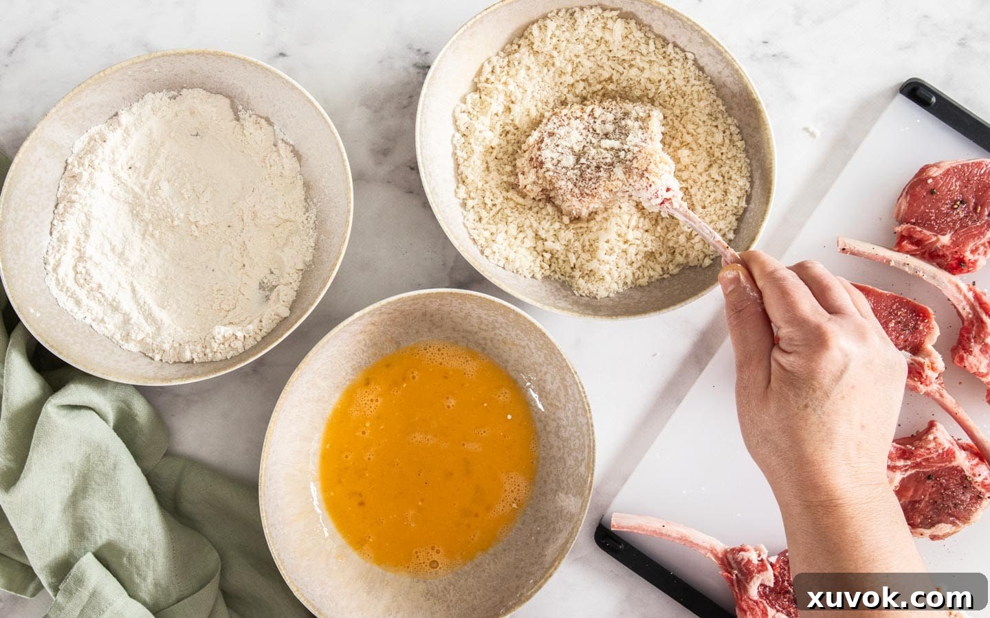 A lamb cutlet being coated in panko breadcrumbs, with bowls of flour and egg visible.