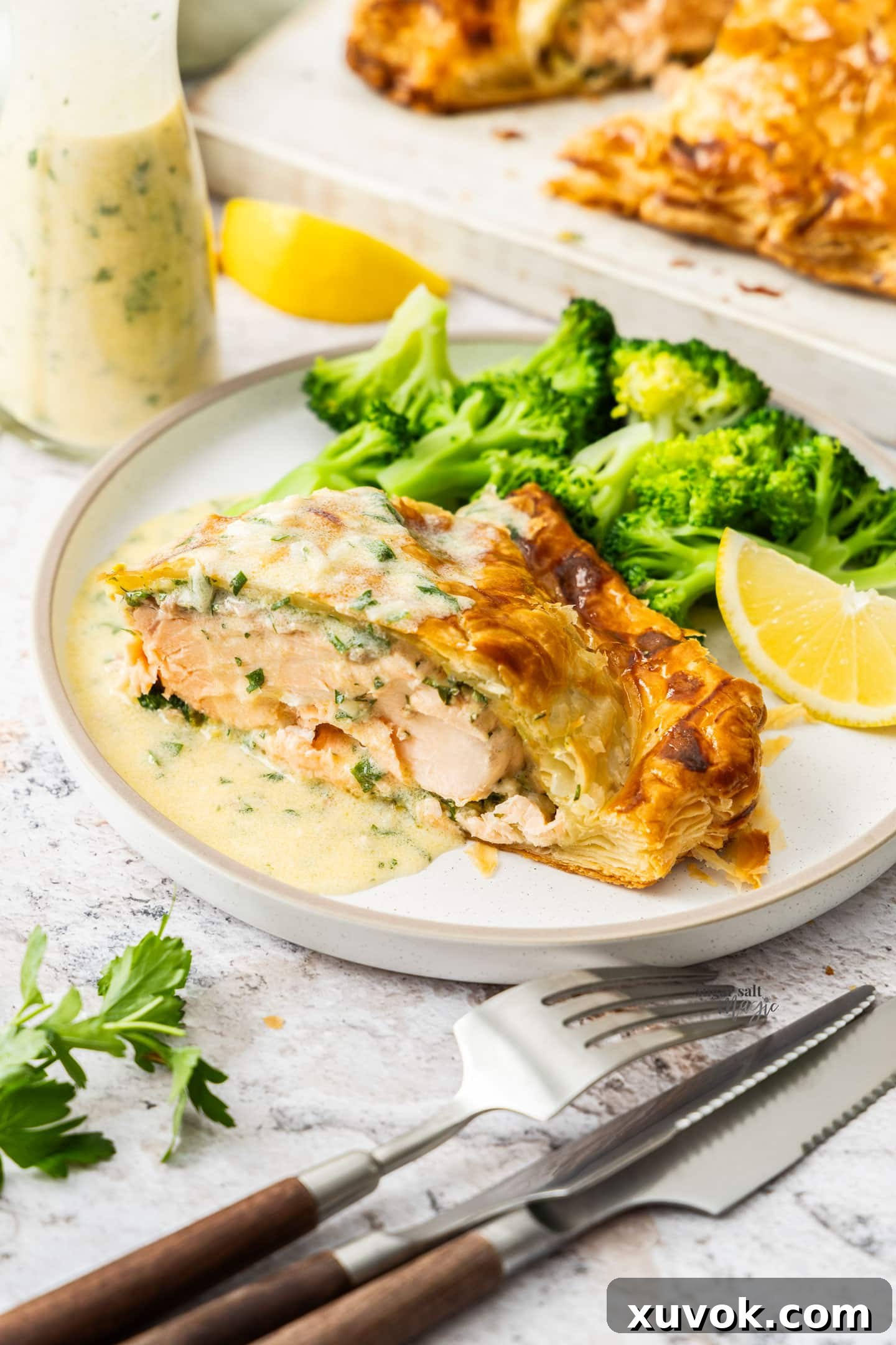 A beautiful slice of salmon wellington on a plate with broccoli and a creamy white wine sauce.