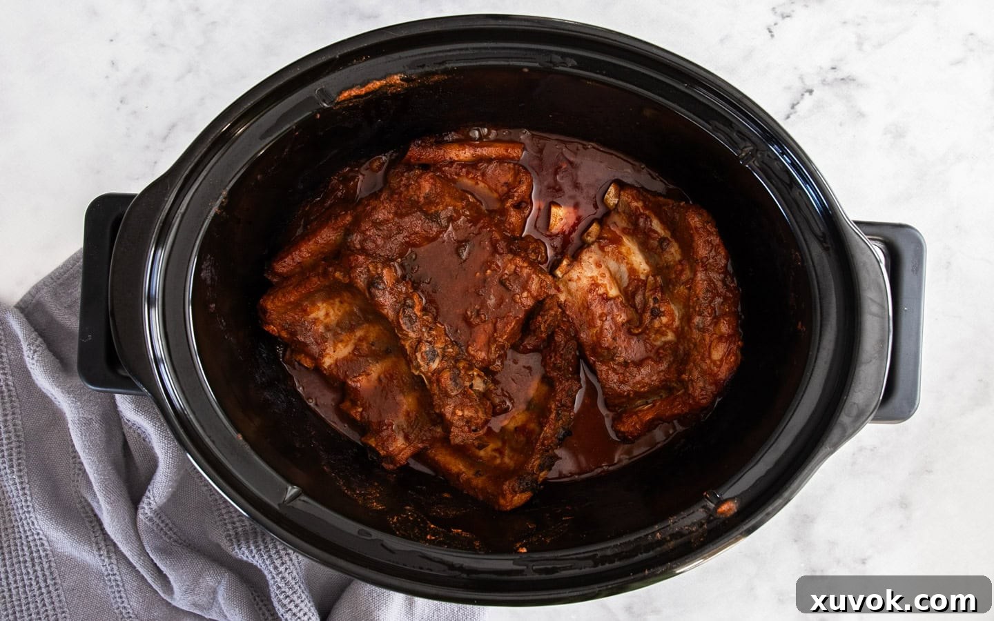 Cooked pork ribs, glistening with BBQ sauce, resting in the slow cooker bowl after tenderizing.