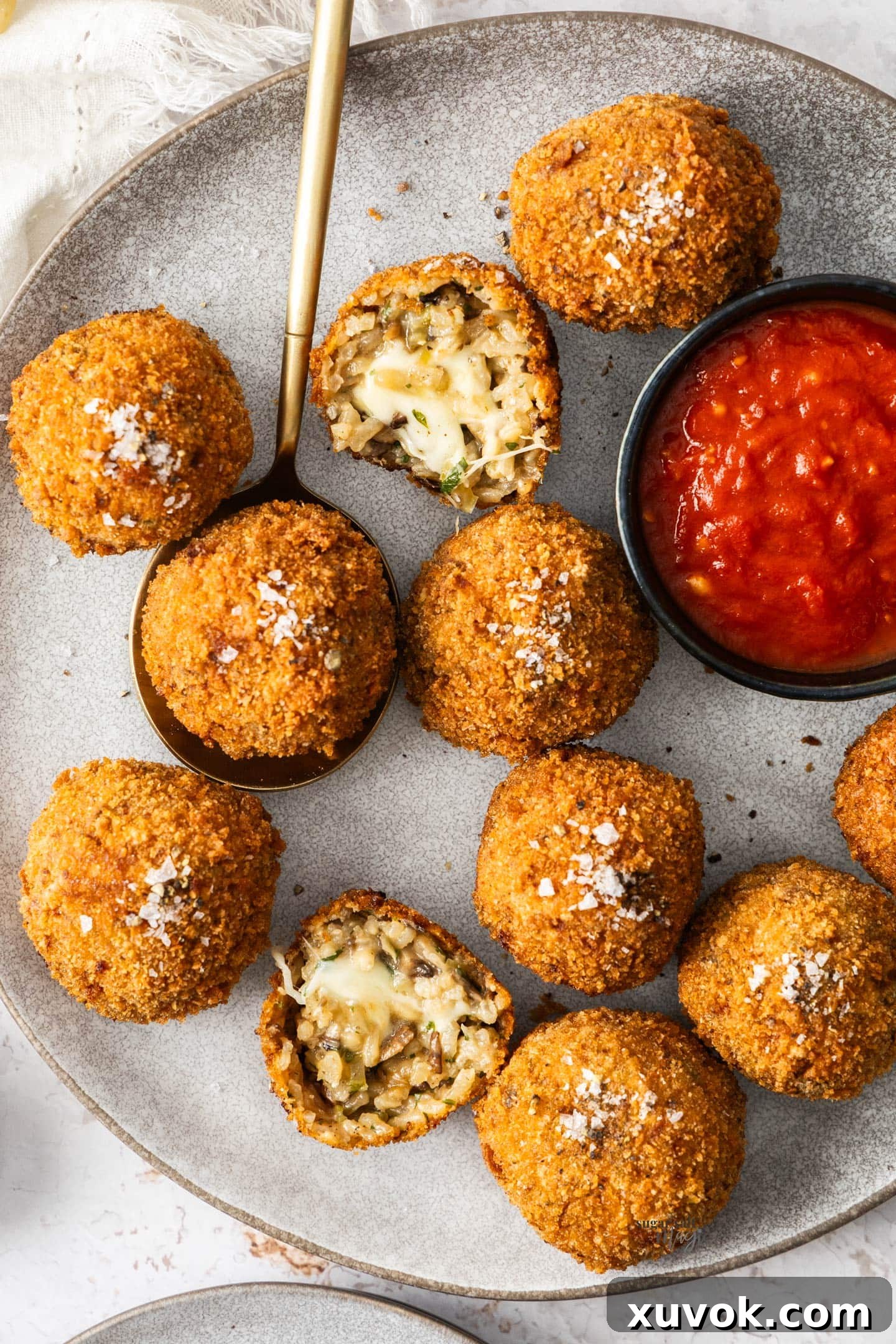 A grey plate featuring ten golden-fried mushroom arancini, with one sliced in half to reveal the creamy risotto and melted mozzarella filling.