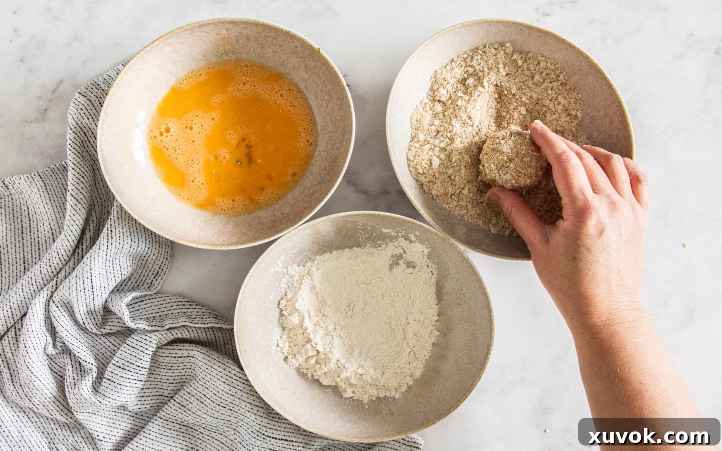 Three shallow bowls arranged for the breading process, containing flour, whisked egg, and breadcrumbs, with an arancini ball being rolled in the breadcrumbs.