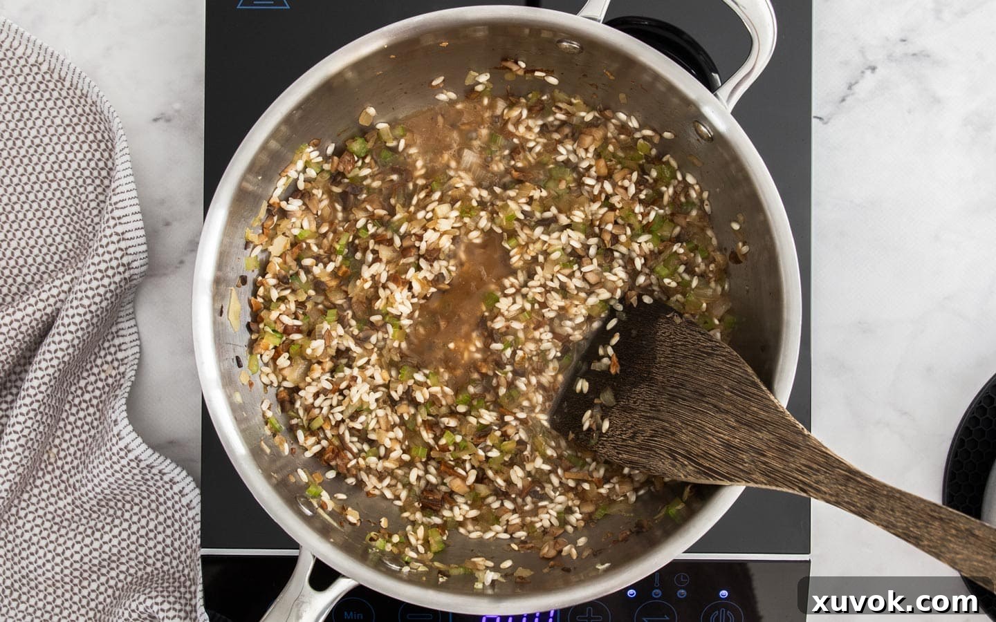 A ladle adding a portion of hot stock to the rice and vegetable mixture in a pan, steam gently rising.