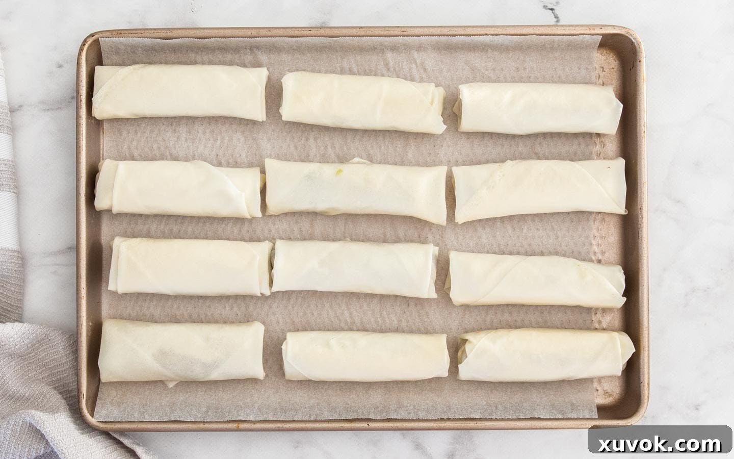 12 perfectly rolled prawn spring rolls arranged on a baking tray, ready for frying.