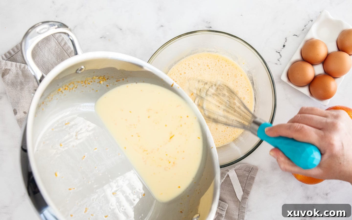 Hot orange-infused cream being slowly poured into a bowl of whisked egg yolks, tempering the mixture.