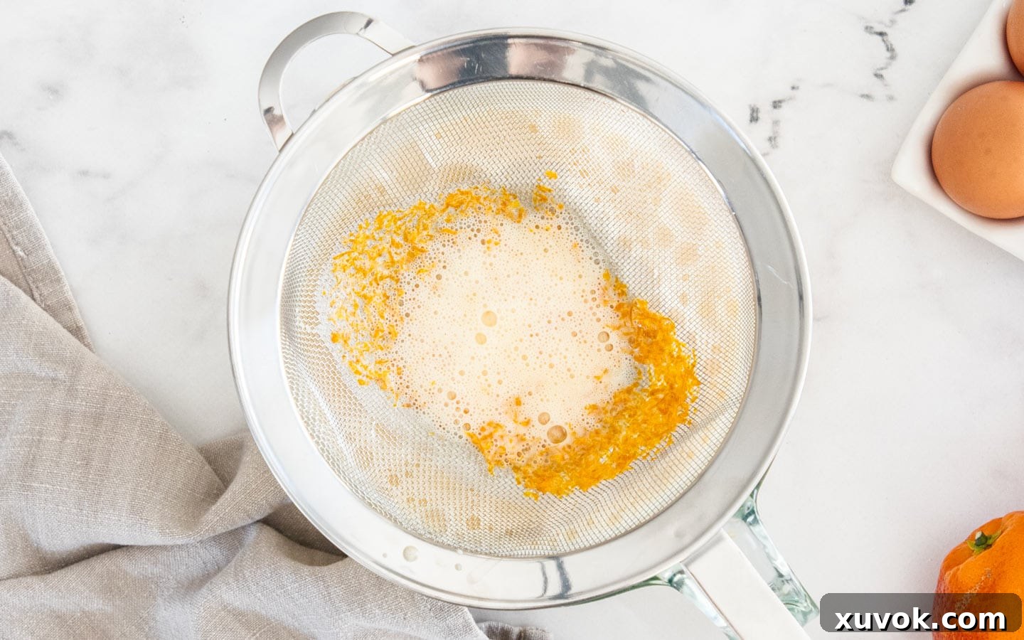 Orange custard mixture being strained through a fine-mesh sieve into a clean bowl, ensuring a smooth texture.