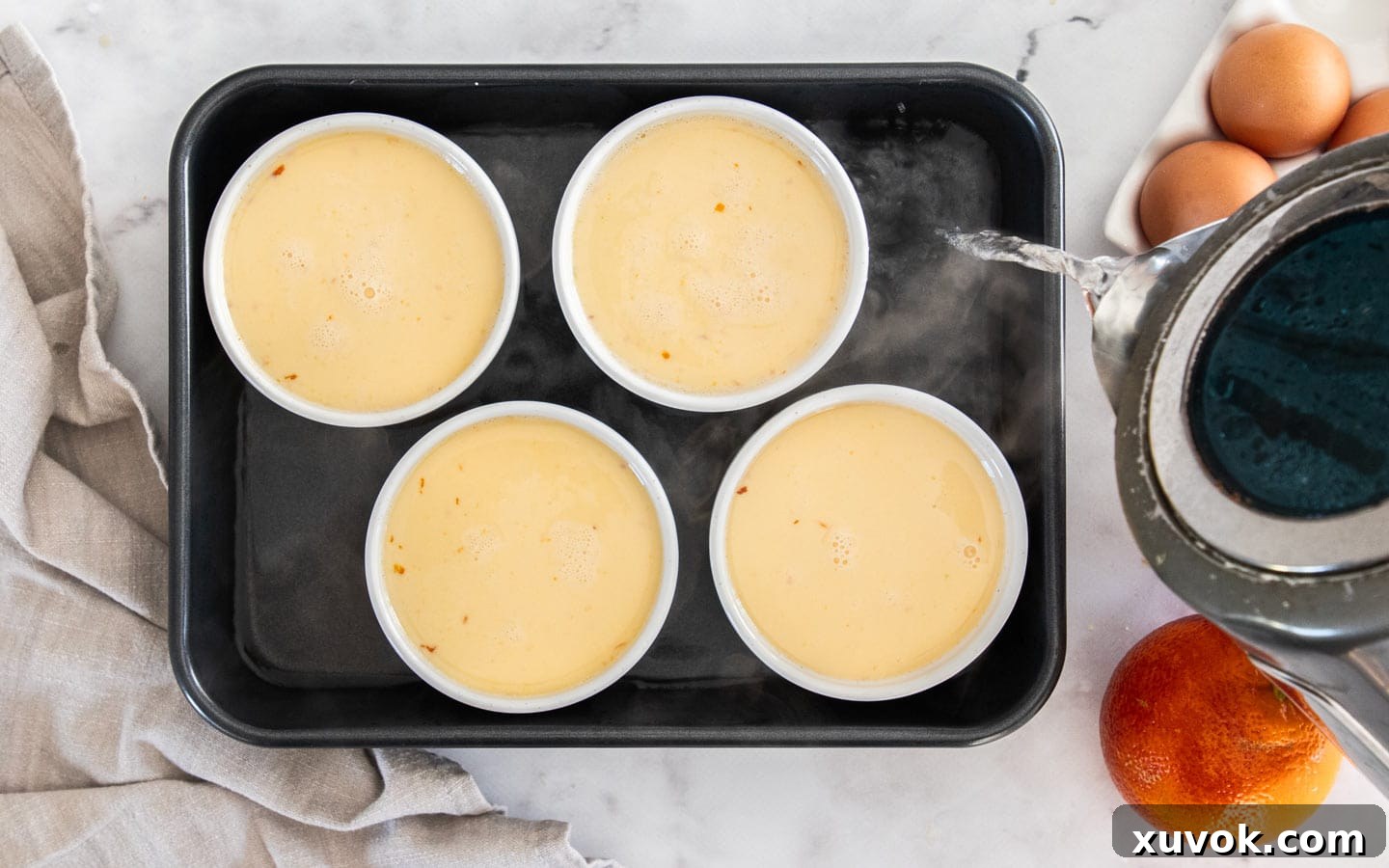 Hot water being poured into a baking tray around ramekins filled with orange custard, creating a water bath.