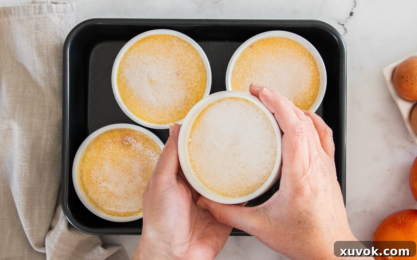 Hands shaking a ramekin to evenly distribute sugar over the top of the chilled orange creme brulees before torching.