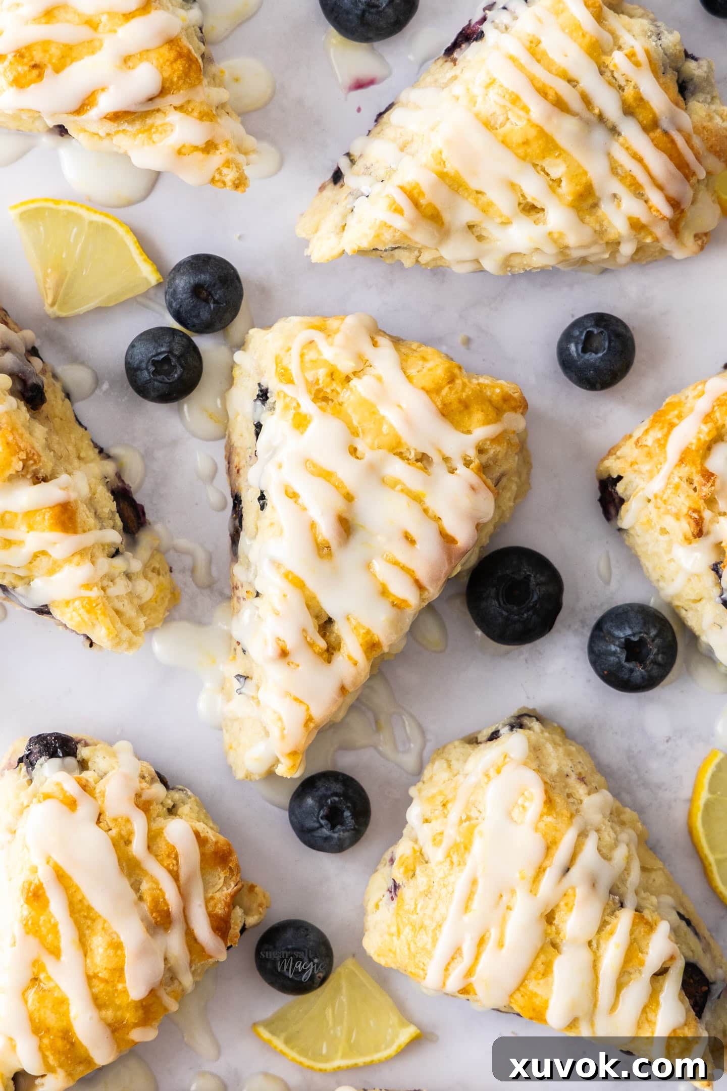 Top down view of 7 triangle blueberry and lemon scones with lemon glaze, surrounded by fresh blueberries and lemon slices.