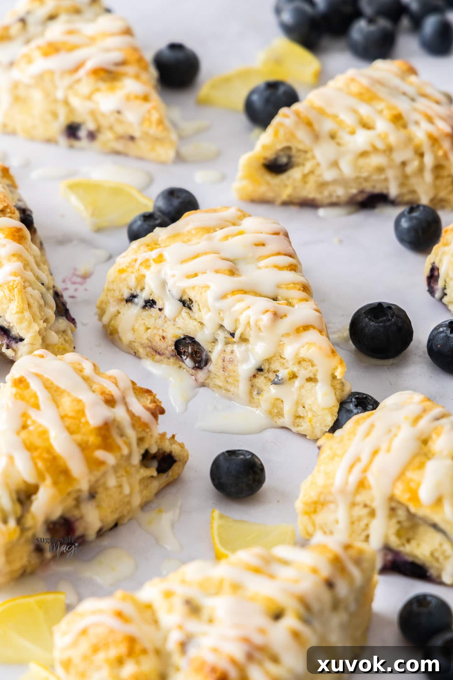 Focus on a central triangular scone studded with blueberries and a lemon glaze on top, with other scones in the background.