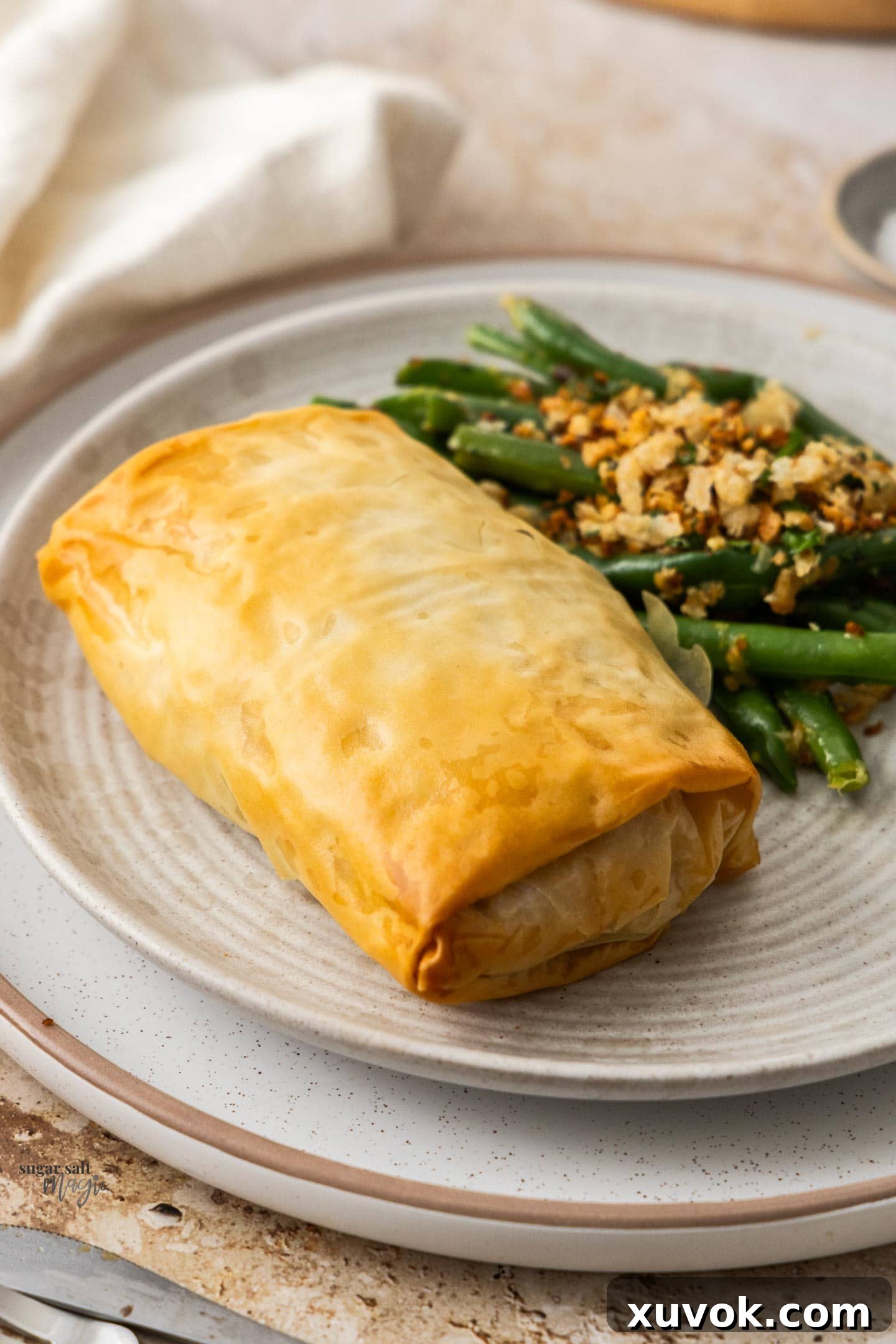 A perfectly baked chicken filo parcel, glistening golden brown, served on a plate with fresh green beans in the background, highlighting a complete meal.