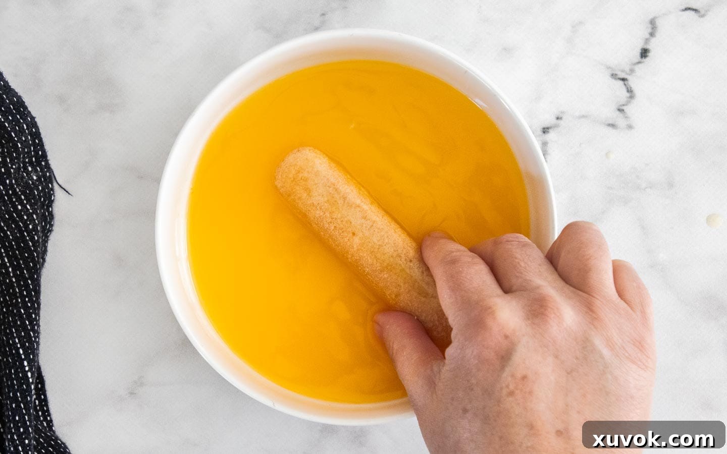 A hand gently dunking a Savoiardi biscuit into a bowl of vibrant orange juice, ready for the tiramisu layers.