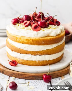 A white forest cake, 3 layers of cake and cream, topped with cherries, on a platter.