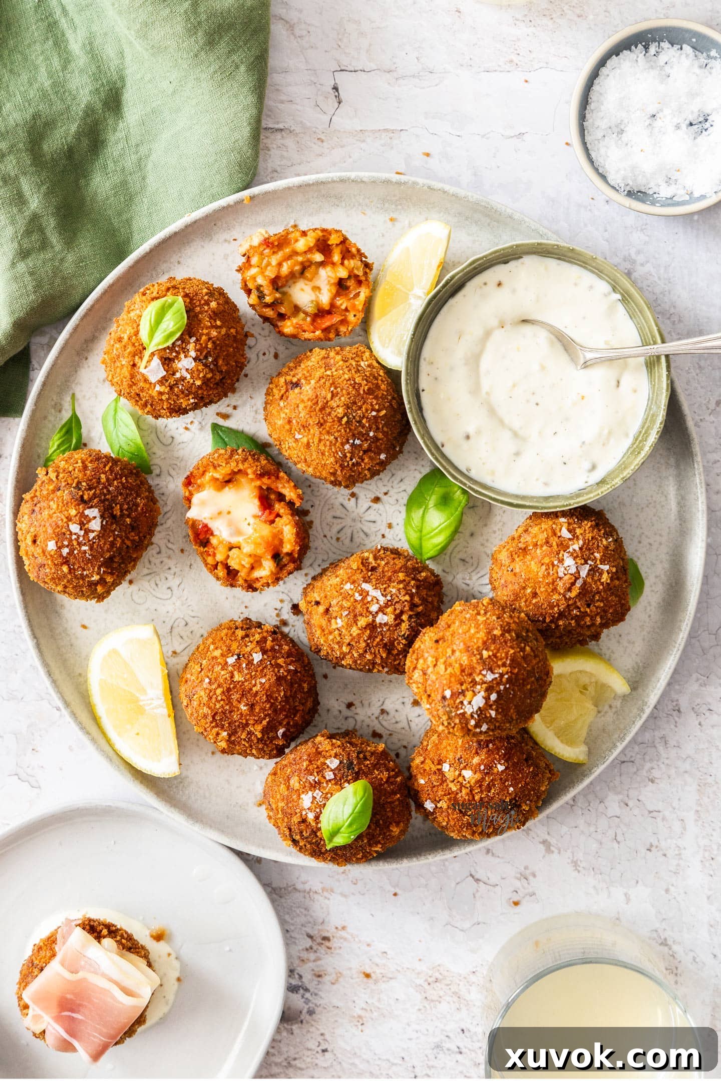 Top-down view of a platter of ten golden-fried tomato arancini, with one broken in half to reveal the molten mozzarella cheese inside. Lemon wedges are scattered around.