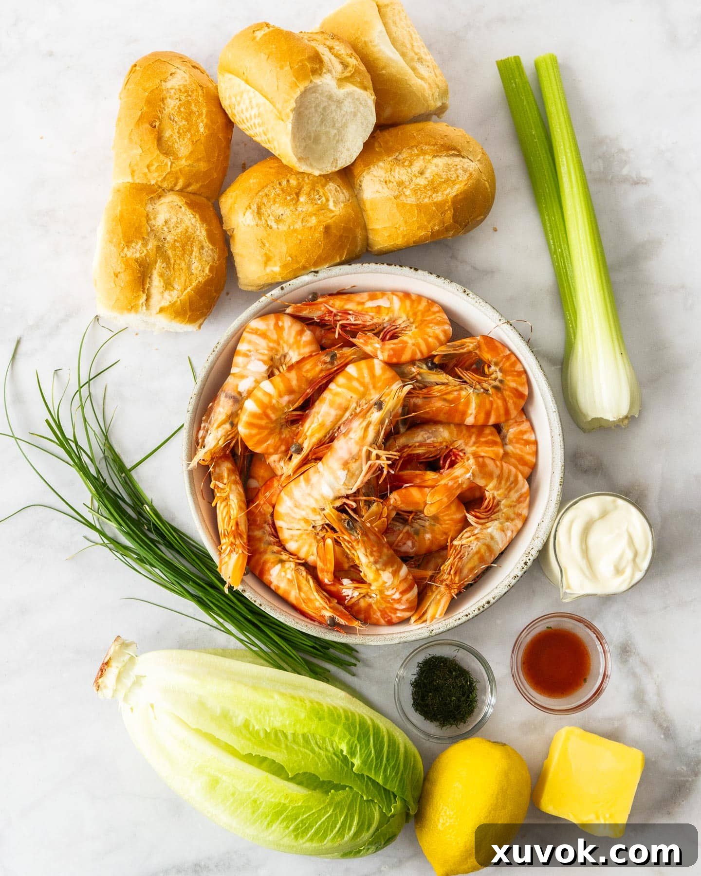 Fresh ingredients laid out on a clean marble surface, including prawns, celery, lettuce, lemons, and herbs, all ready for making prawn rolls.