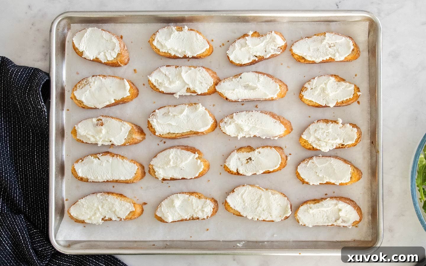 The crostini on a baking sheet spread with goats cheese.