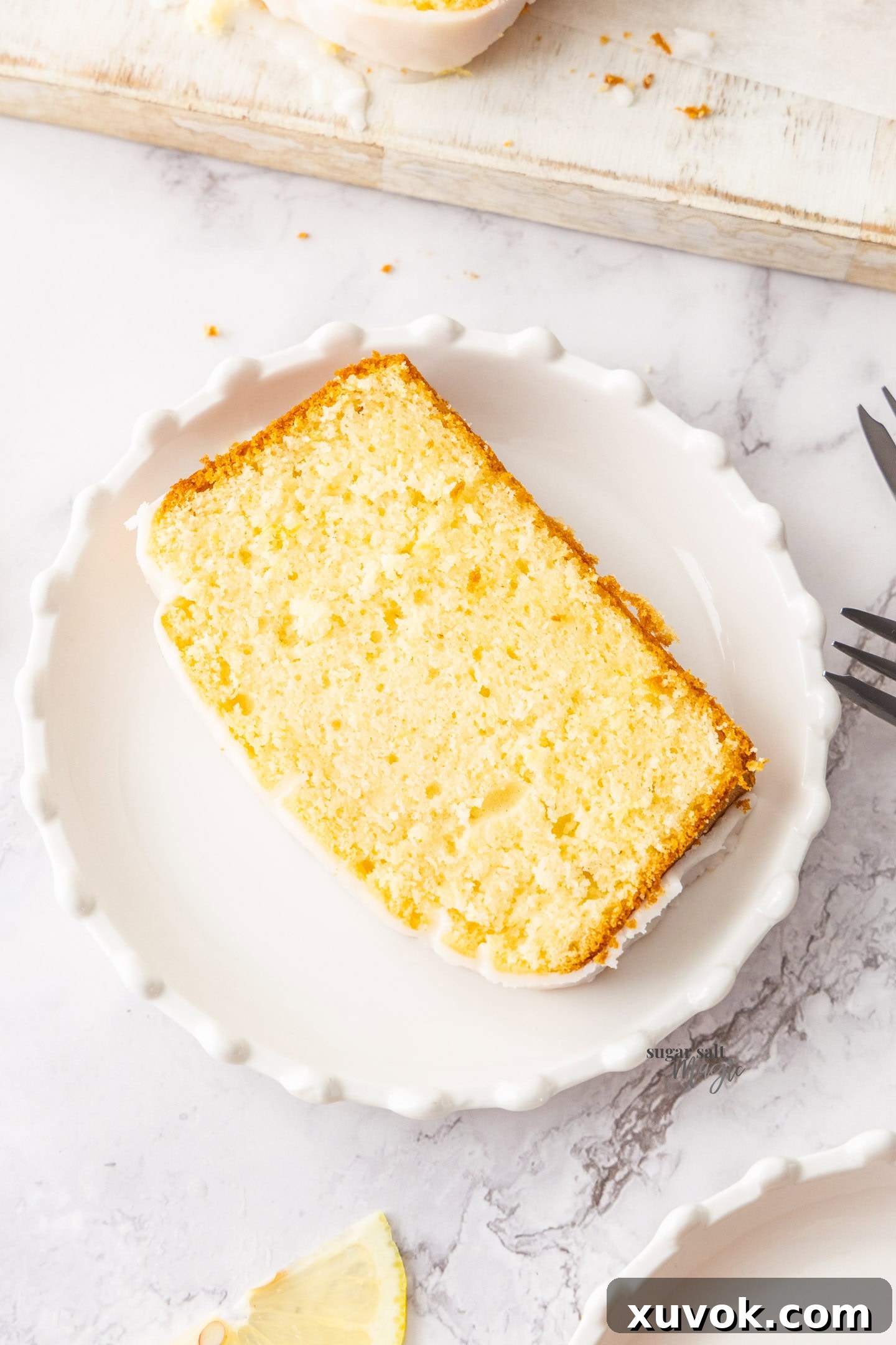A close-up, top-down view of a single slice of lemon loaf cake resting on a small dessert plate, garnished with a light lemon glaze and a delicate lemon zest curl.