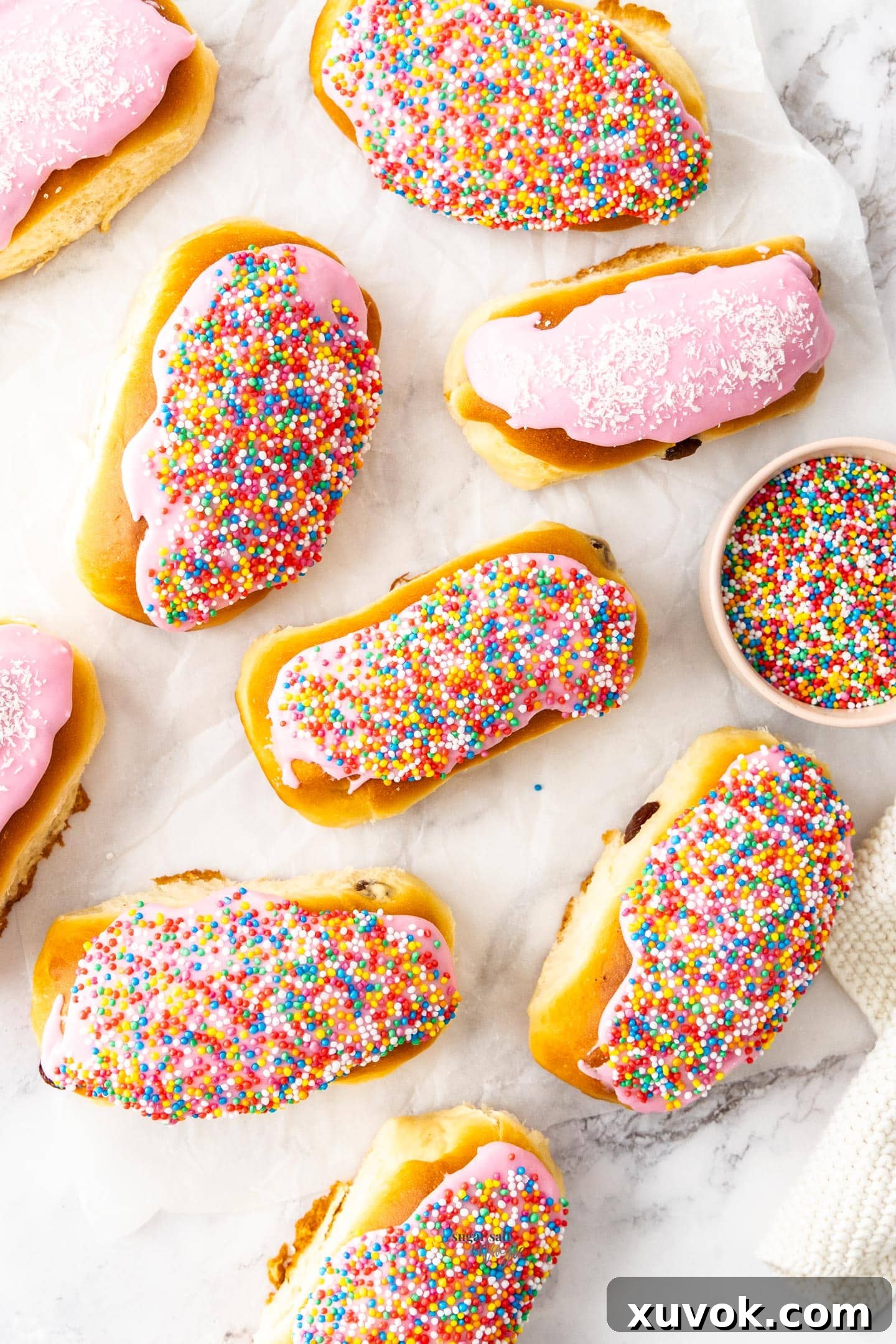 Top down view of 9 freshly baked finger buns, generously topped with pink icing and colorful 100s and 1000s, arranged on a sheet of baking paper, evoking a sense of classic Australian bakery treats.