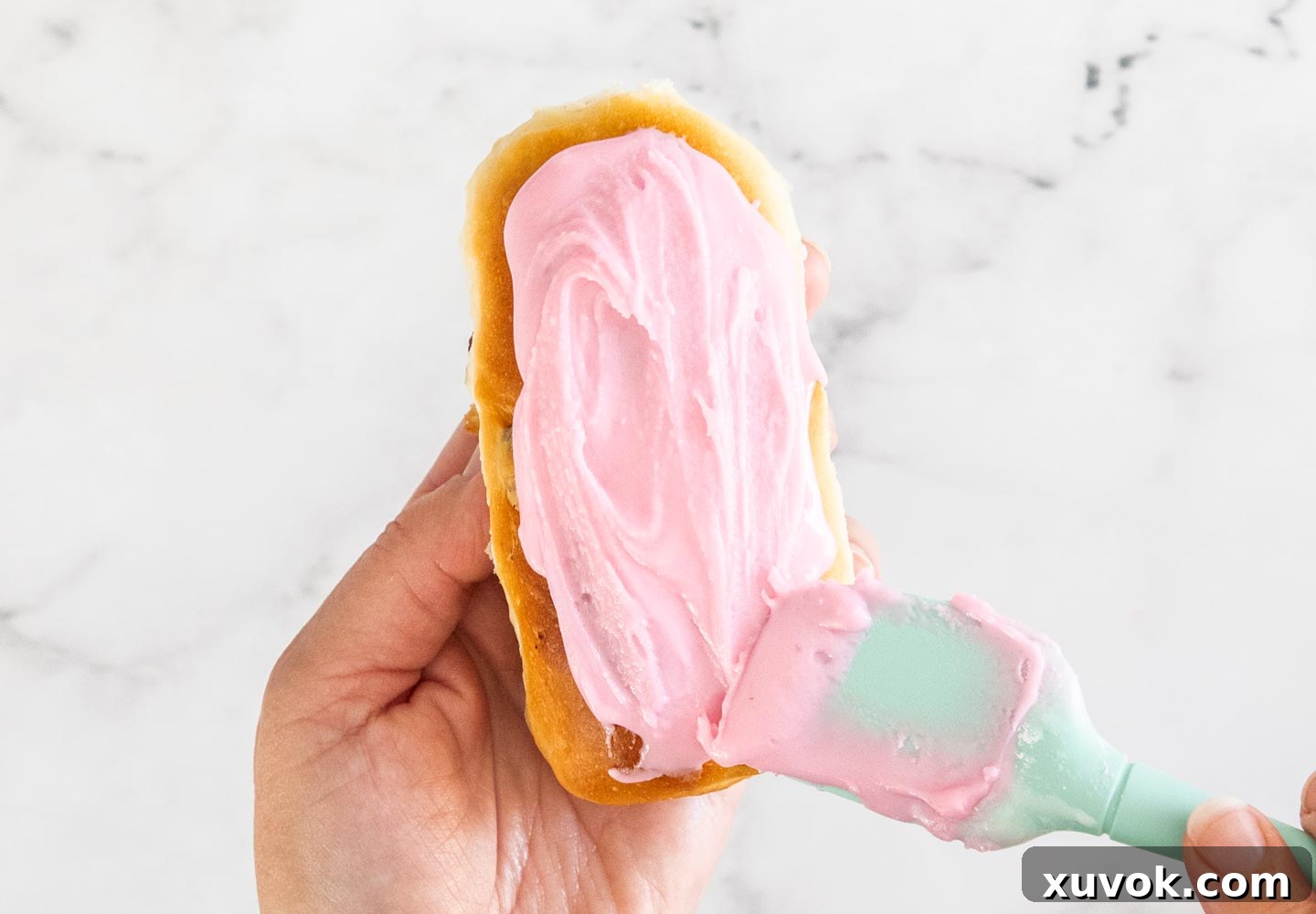 A spatula smoothly spreading vibrant pink icing onto a freshly baked finger bun, preparing it for its final decorative touch.