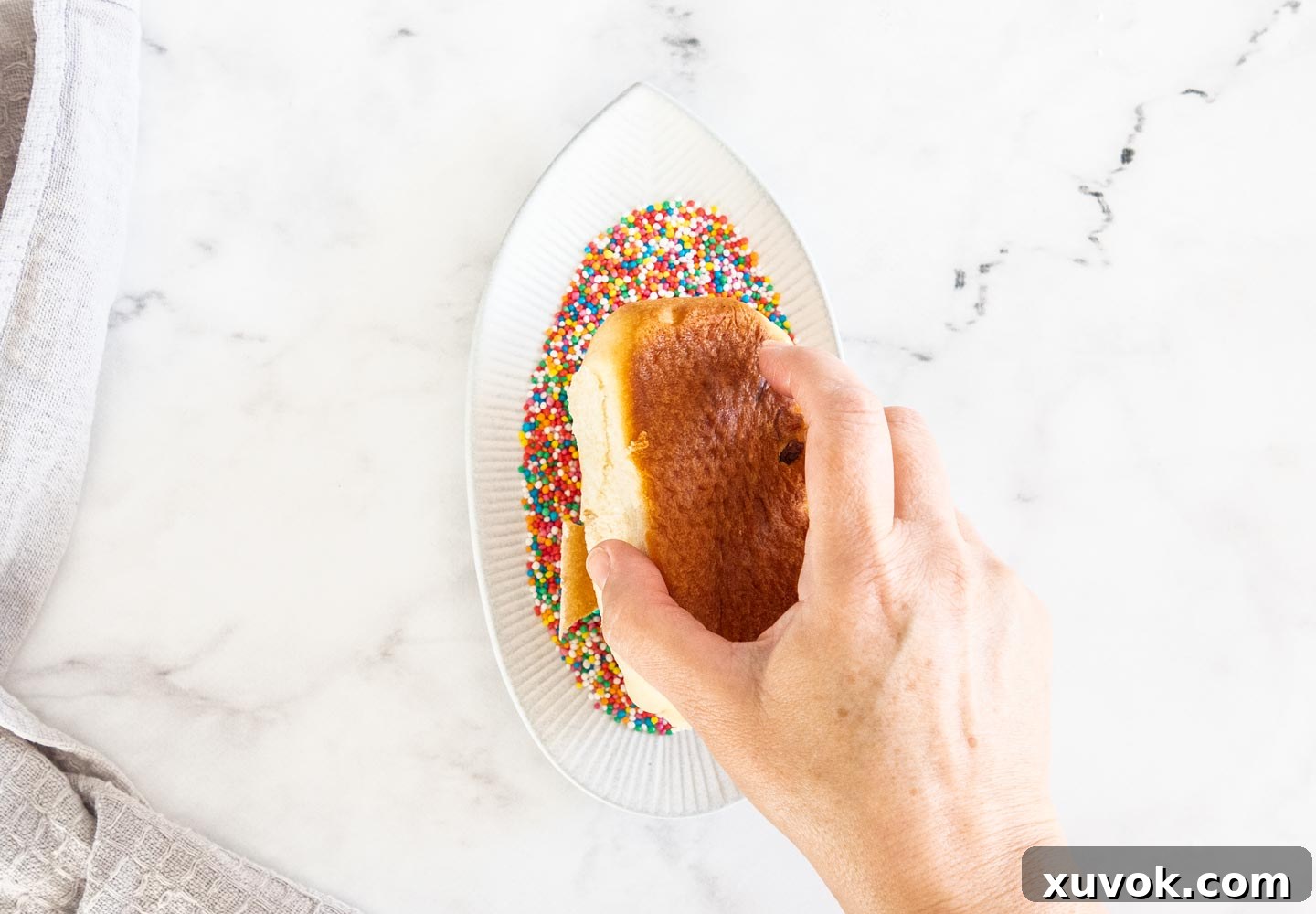 A hand gently pressing the iced top of a finger bun into a bowl of colorful 100s and 1000s, adding the final, classic decoration.