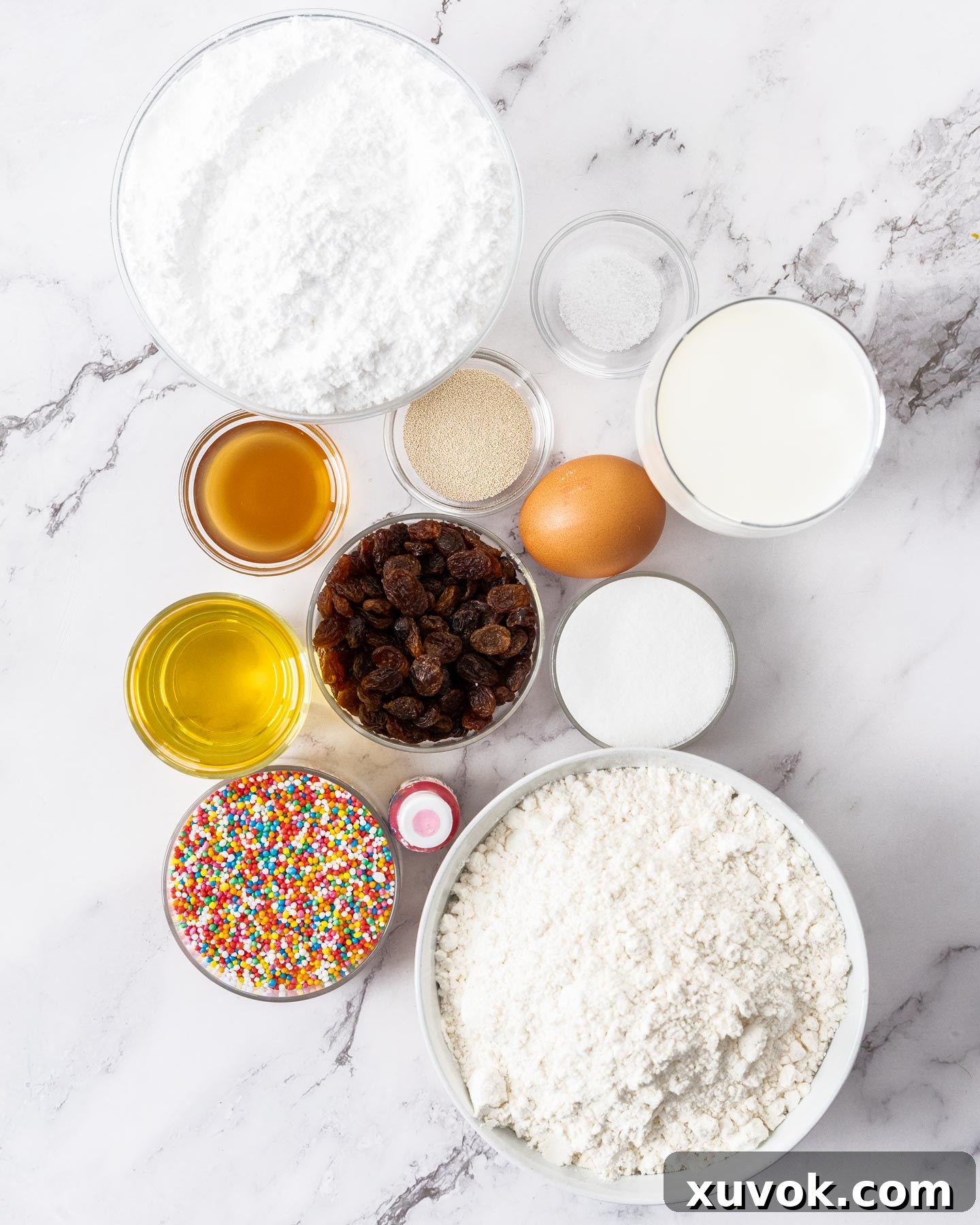 Various finger bun ingredients laid out on a clean marble surface, including flour, sugar, yeast, milk, eggs, and oil, ready for baking.