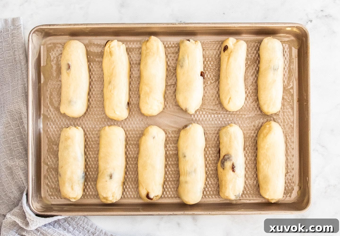 Twelve neatly shaped logs of finger bun dough, evenly spaced on a baking tray, ready for their second proving.