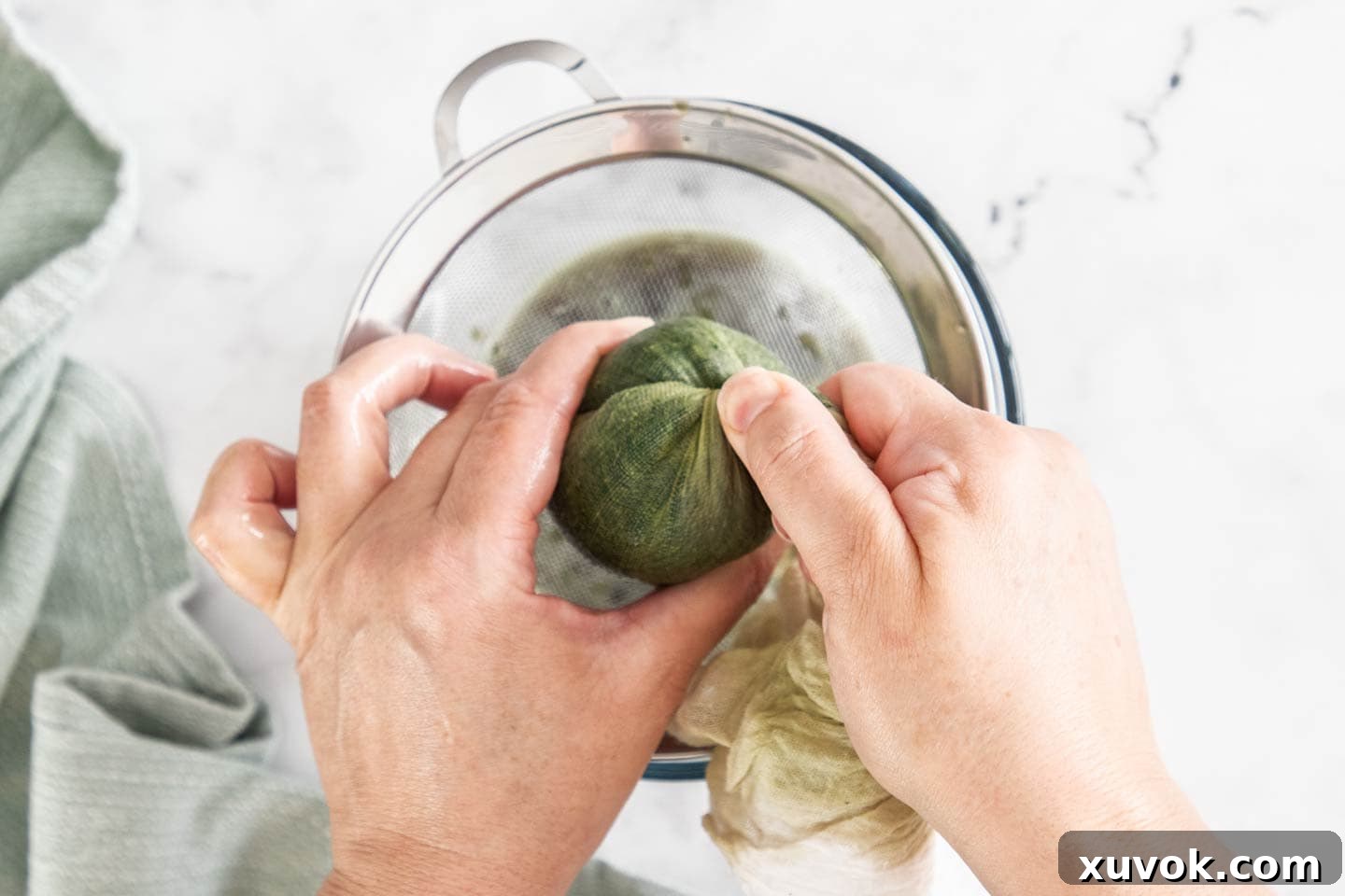 Two hands holding and twisting cheesecloth to squeeze liquid from thawed spinach.
