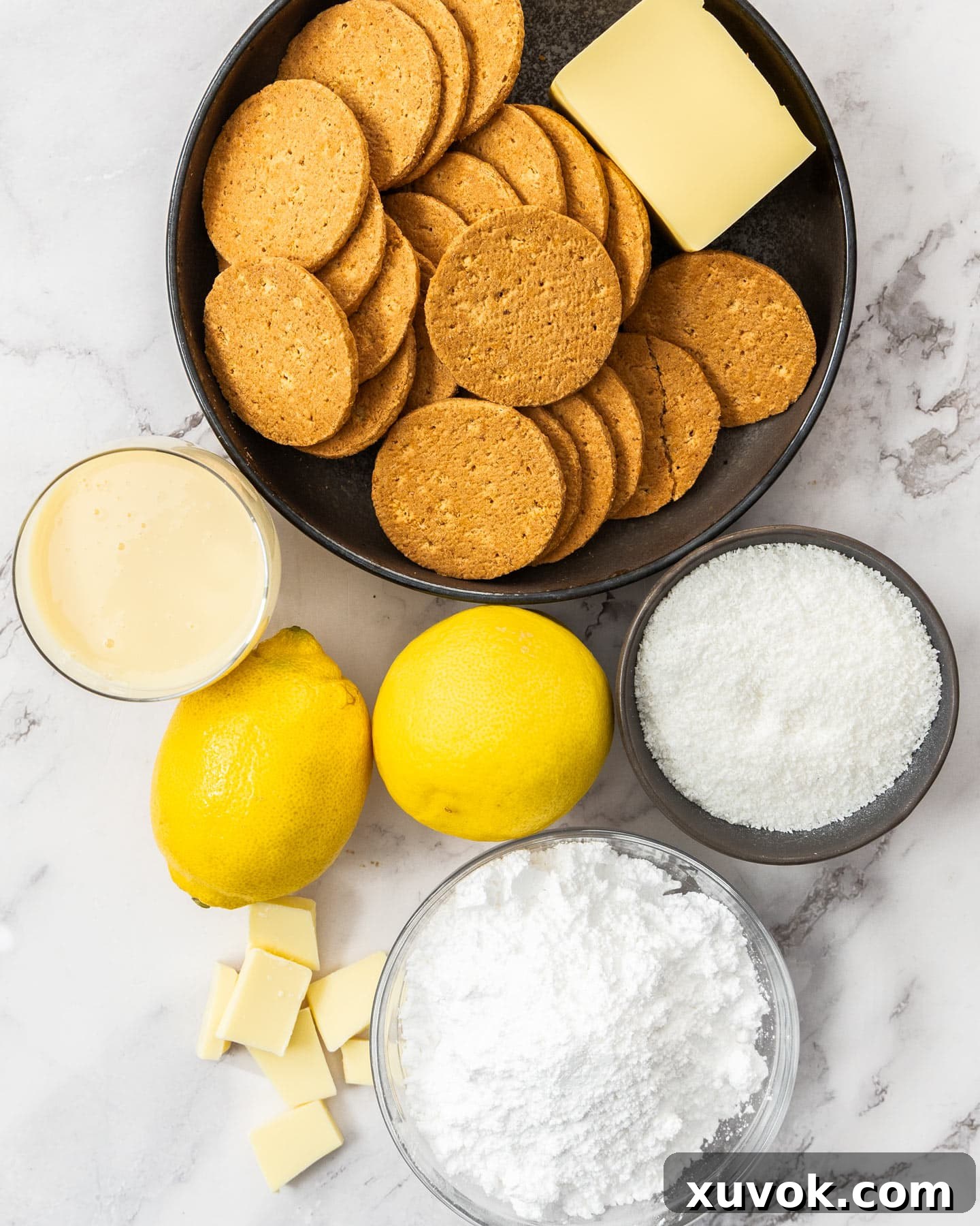 Ingredients for lemon coconut slice laid out on a surface, including lemons, butter, condensed milk, biscuits, and coconut.