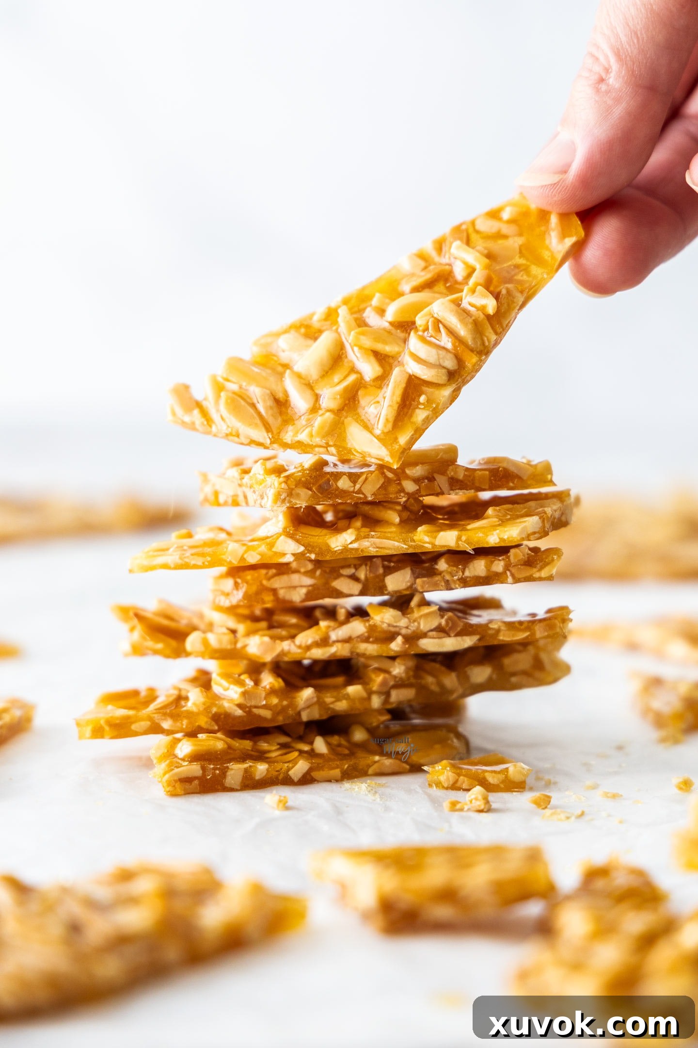 A hand lifting a shard of almond praline off of a stack, showcasing its crunchy texture and golden color.