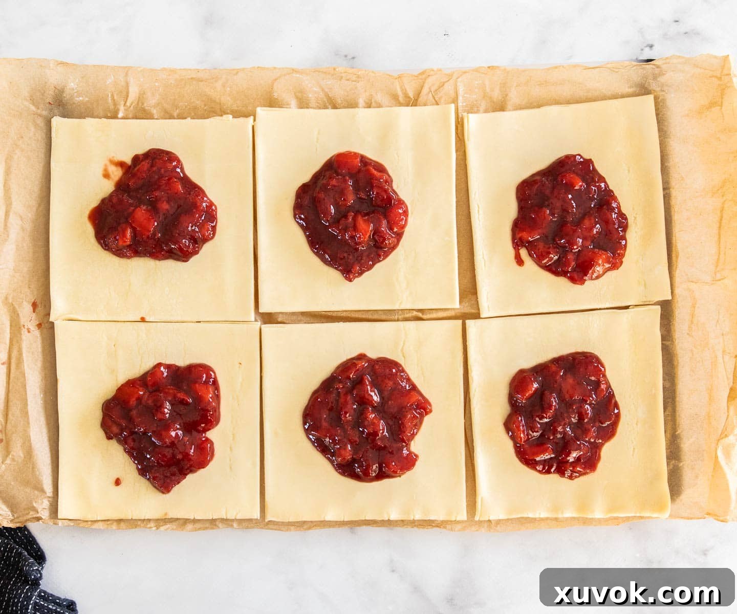 Six small squares of puff pastry neatly arranged, each carefully topped with a spoonful of bright red strawberry filling, ready for folding.