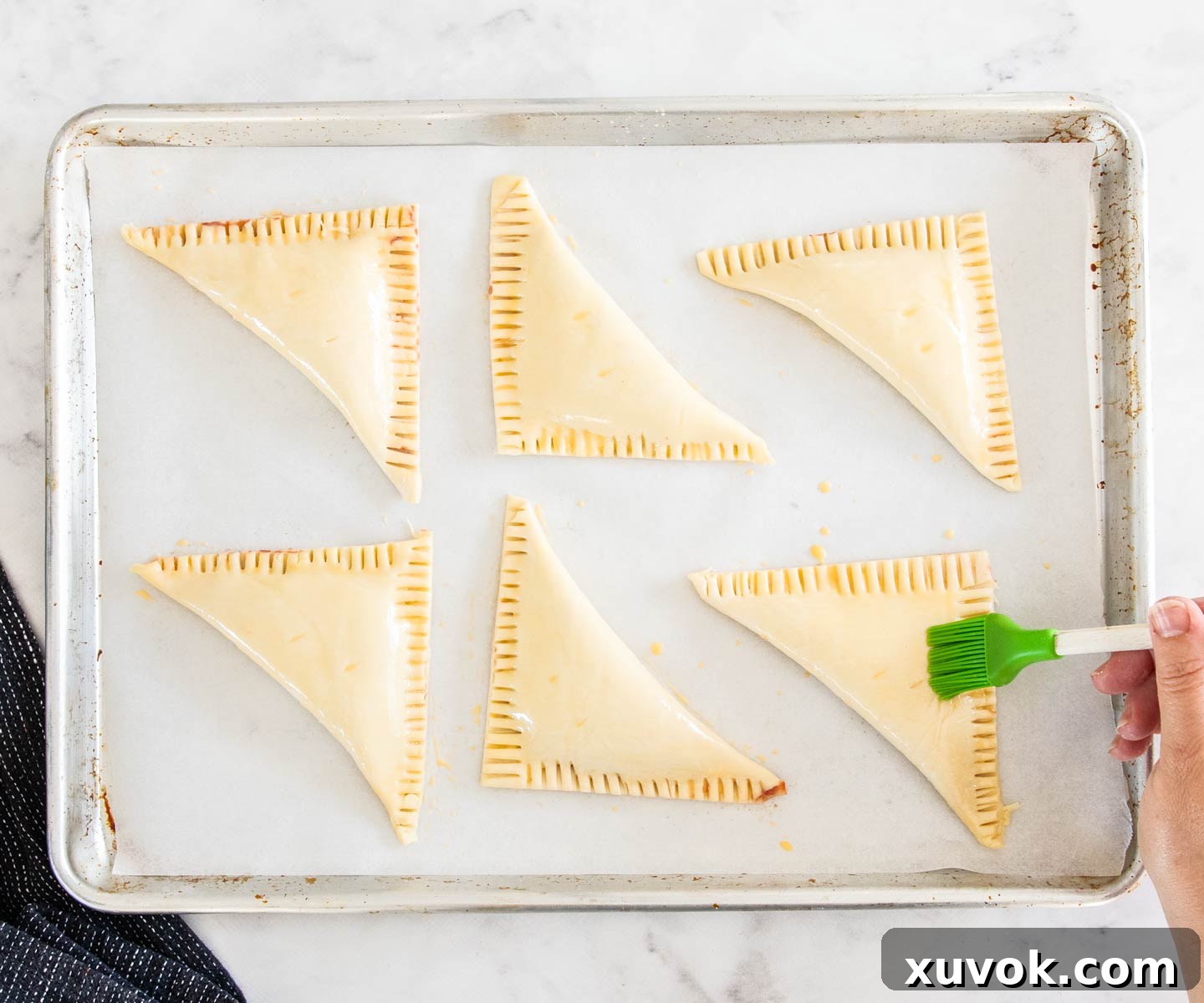 A hand expertly brushing a golden egg wash over the uncooked strawberry turnovers arranged on a baking sheet, preparing them for a perfect bake.