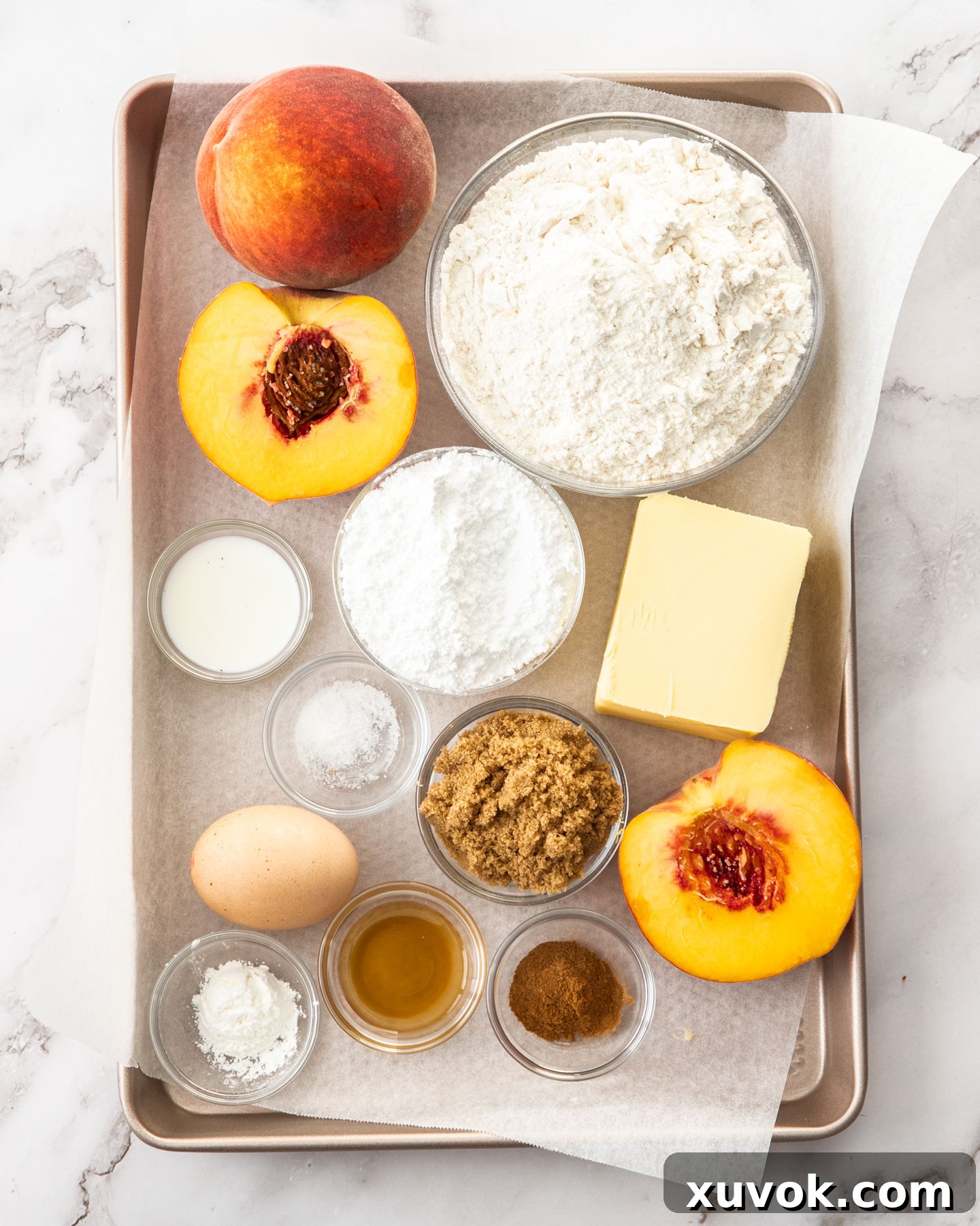A collection of fresh ingredients for peach hand pies, including flour, sugar, butter, ripe peaches, cinnamon, and vanilla, arranged neatly on a baking tray.